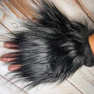 Peut inclure: Un manchon de poignet en fausse fourrure noire avec de longues fibres hirsutes. Le manchon est enroulé autour d'un poignet, avec les doigts d'une main visibles. La fourrure a un aspect sombre et texturé, suggérant un costume ou un accessoire.
