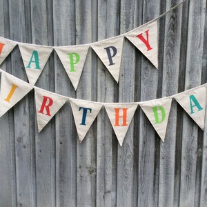 May include: A white fabric banner with the words "Happy Birthday" in colorful letters. The banner is strung up on a wooden fence.