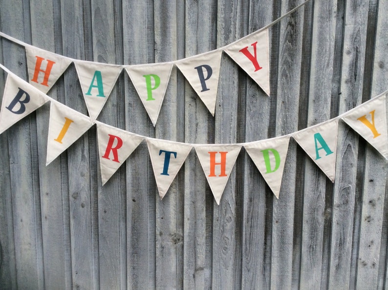 May include: A white fabric banner with the words "Happy Birthday" in colorful letters. The banner is strung up on a wooden fence.