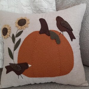 Peut inclure: Un coussin carré beige orné d'une citrouille orange appliquée au centre, de deux oiseaux marron perchés sur la citrouille, et de deux tournesols jaunes avec des tiges et des feuilles vert foncé sur le côté gauche, le tout en feutrine.