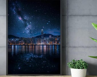 以下が含まれることがあります： 星空の下の夜の都市景観を描いた額入りのプリント。画像は、水面に映る都市のスカイラインの上に、星でいっぱいの濃い青色の空を示しています。プリントは木製のテーブルに飾られています。