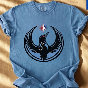May include: A blue jean colored t-shirt featuring a black bird design with a red eye and a star. The bird is encircled by a black and blue design. The shirt has rolled-up sleeves and the text "Blue Jean" is at the bottom.