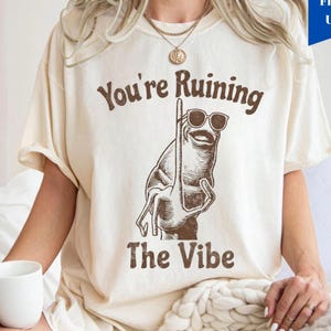 Puede incluir: Camiseta color marfil con un gráfico marrón de un personaje de dibujos animados con gafas de sol y un palo. El texto "You're Ruining The Vibe" está impreso en marrón arriba y abajo del gráfico. La camiseta es de corte holgado.