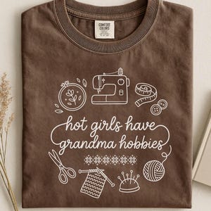 May include: Espresso-coloured t-shirt with a white graphic of sewing and knitting tools, and the phrase "hot girls have grandma hobbies." The shirt is folded and has a Comfort Colors label.
