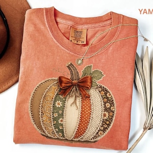May include: A garment-dyed orange t-shirt featuring a graphic of a patchwork pumpkin with a brown velvet bow. The pumpkin design includes various floral and textured patterns in shades of green, orange, and cream. The shirt has a crew neckline.