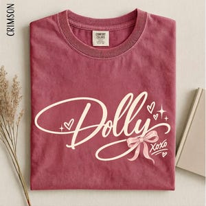 May include: A crimson short-sleeve t-shirt featuring the word Dolly in a large, cream-colored cursive script. The design includes a small cream-colored bow, several hearts, and star accents, with the text XOXO printed in the lower right corner.