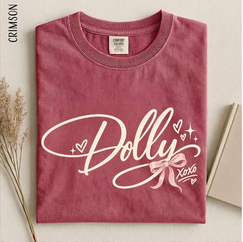 May include: A crimson short-sleeve t-shirt featuring the word Dolly in a large, cream-colored cursive script. The design includes a small cream-colored bow, several hearts, and star accents, with the text XOXO printed in the lower right corner.