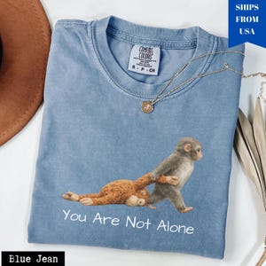 Puede incluir: Camiseta azul vaquero Comfort Colors con un gr&aacute;fico de monos y el texto "You Are Not Alone". Un collar dorado est&aacute; sobre la camiseta. La camiseta es de color azul claro.