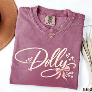 May include: A berry-colored short-sleeve t-shirt featuring the word Dolly in a cream-colored script font. The design includes small hearts, starbursts, a ribbon bow, and the text XOXO. The shirt has a Comfort Colors brand label at the neck.