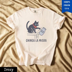 May include: Ivory-colored t-shirt featuring a cartoon cat wearing a red bandana, reaching for a gray ice jug. The text "CHINGA LA MIGRA" is printed below the cat. The shirt is made of soft material.