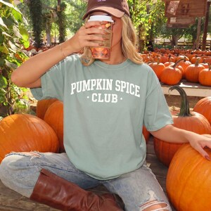 May include: A sage green short-sleeve t-shirt featuring the white text PUMPKIN SPICE CLUB printed in a collegiate font on the chest. The shirt is worn with light blue distressed denim jeans and brown leather boots in a pumpkin patch setting.