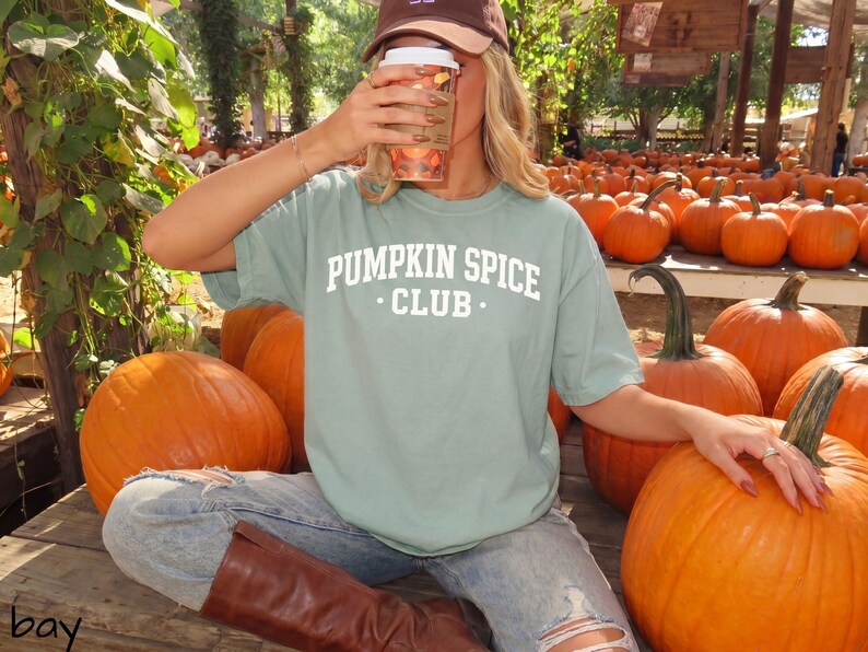 May include: A sage green short-sleeve t-shirt featuring the white text PUMPKIN SPICE CLUB printed in a collegiate font on the chest. The shirt is worn with light blue distressed denim jeans and brown leather boots in a pumpkin patch setting.