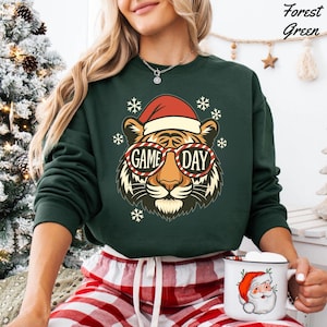 May include: Forest green sweatshirt featuring a tiger wearing a Santa hat and sunglasses with "GAME DAY" text. The design includes snowflakes. The model is wearing red and white plaid pajama pants and holding a mug with a Santa Claus design.