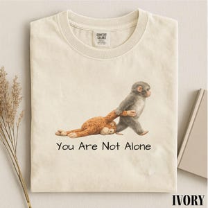 Puede incluir: Camiseta color marfil con un gr&aacute;fico de dos monos. Un mono gris tira de un mono marr&oacute;n, con el texto "You Are Not Alone" debajo. La camiseta tiene una etiqueta Comfort Colors.