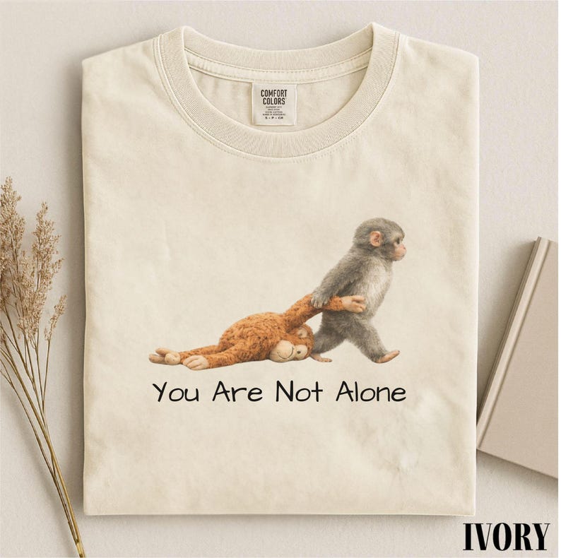 Puede incluir: Camiseta color marfil con un gr&aacute;fico de dos monos. Un mono gris tira de un mono marr&oacute;n, con el texto "You Are Not Alone" debajo. La camiseta tiene una etiqueta Comfort Colors.
