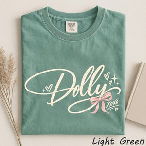 May include: A sage green short-sleeve t-shirt featuring the word Dolly in a white cursive script. The design includes a light pink bow, small white hearts, and the text XOXO. The shirt has a crew neckline and a Comfort Colors brand label.