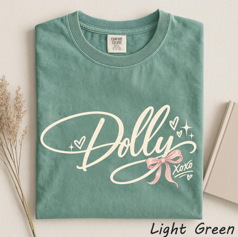 May include: A sage green short-sleeve t-shirt featuring the word Dolly in a white cursive script. The design includes a light pink bow, small white hearts, and the text XOXO. The shirt has a crew neckline and a Comfort Colors brand label.