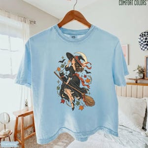 May include: A light blue short-sleeve t-shirt featuring a graphic illustration of a witch in a black dress riding a broomstick with a black cat, a crescent moon, and scattered autumn leaves in shades of orange, red, and green.