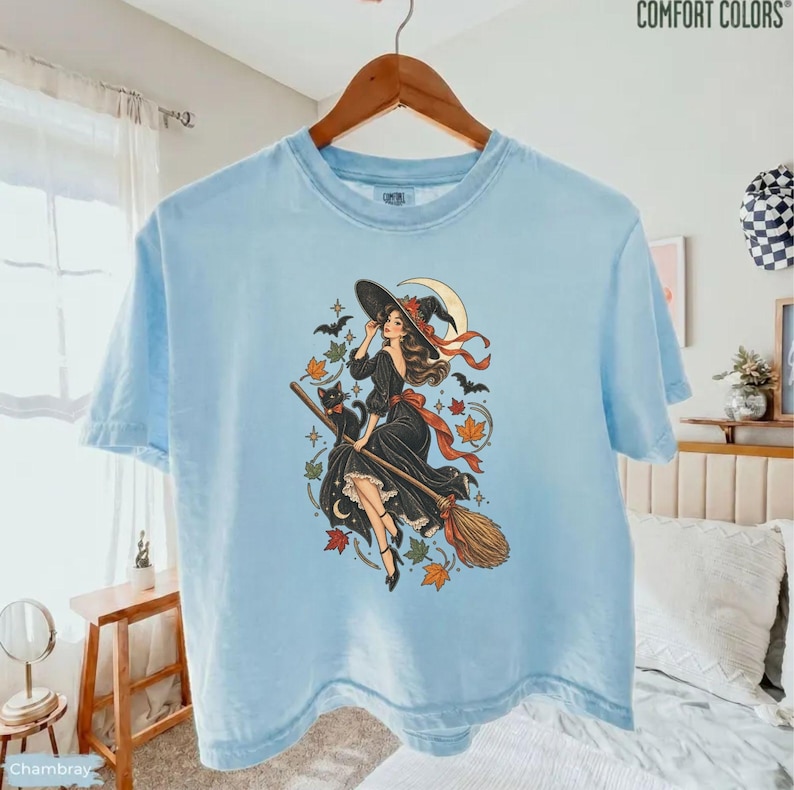 May include: A light blue short-sleeve t-shirt featuring a graphic illustration of a witch in a black dress riding a broomstick with a black cat, a crescent moon, and scattered autumn leaves in shades of orange, red, and green.
