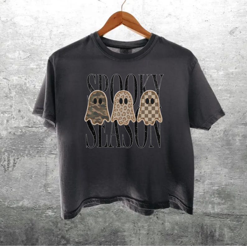 May include: A charcoal gray short-sleeve t-shirt featuring a graphic of three ghosts with different patterns: camouflage, leopard print, and a brown checkerboard. The text "SPOOKY SEASON" is printed in a distressed black font behind the ghosts.