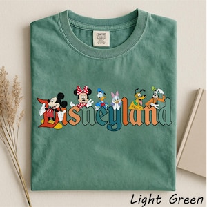 May include: Light green t-shirt featuring the word "Disneyland" in colorful letters with cartoon characters Mickey Mouse, Minnie Mouse, Donald Duck, Daisy Duck, Pluto, and Goofy. The shirt is made of a soft material and has a crew neck.