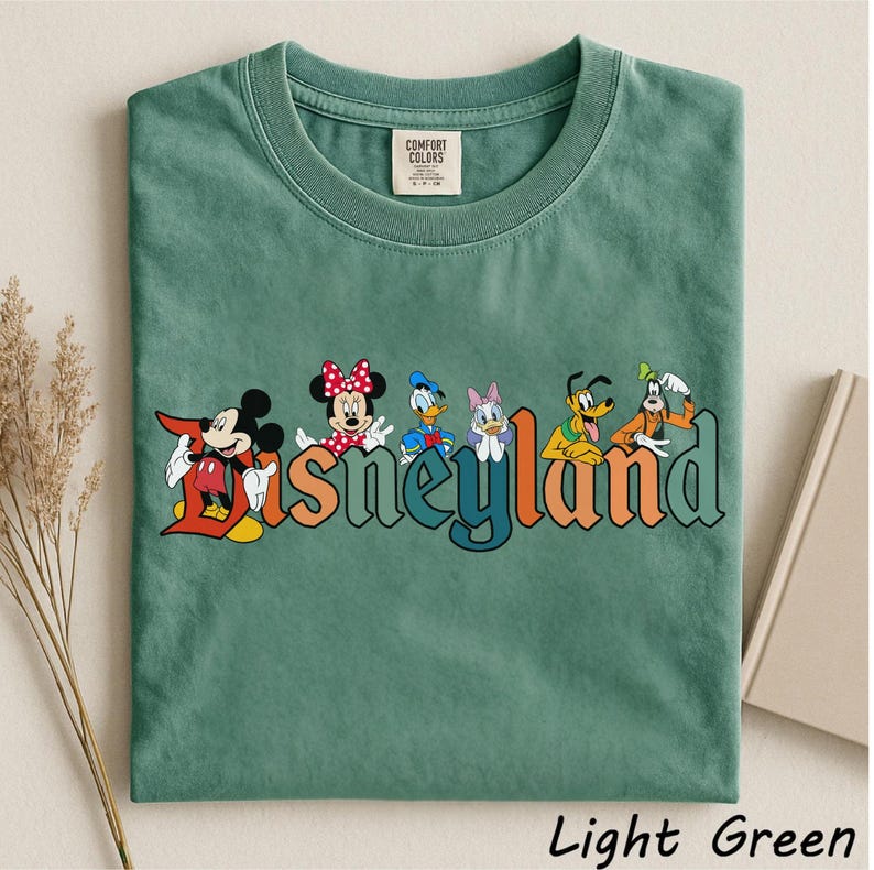 May include: Light green t-shirt featuring the word "Disneyland" in colorful letters with cartoon characters Mickey Mouse, Minnie Mouse, Donald Duck, Daisy Duck, Pluto, and Goofy. The shirt is made of a soft material and has a crew neck.