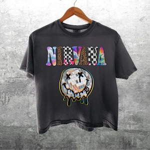May include: A charcoal gray short-sleeve t-shirt featuring the word NIRVANA in multicolored, patterned letters above a graphic of a melting disco ball smiley face with X-shaped eyes and a tongue sticking out, outlined in a bright yellow border.
