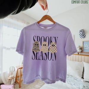May include: A light purple short-sleeve t-shirt with the text "SPOOKY SEASON" in black lettering. The design features three ghost illustrations with different patterns: camouflage, leopard print, and a tan and white checkerboard pattern.