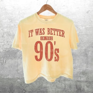 May include: A pale yellow short-sleeved t-shirt featuring distressed red text that reads IT WAS BETTER IN THE 90's. The shirt has a crew neckline and is displayed on a wooden hanger, showcasing a simple design with a vintage-inspired graphic print.