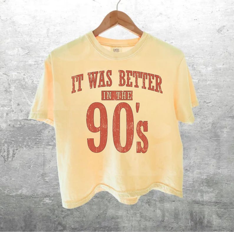 May include: A pale yellow short-sleeved t-shirt featuring distressed red text that reads IT WAS BETTER IN THE 90's. The shirt has a crew neckline and is displayed on a wooden hanger, showcasing a simple design with a vintage-inspired graphic print.