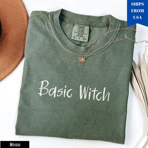 May include: A moss green t-shirt with the phrase "Basic Witch" in white script. The shirt has a crew neck and short sleeves. A gold necklace with a pendant rests on the shirt. The shirt is folded on a white surface.