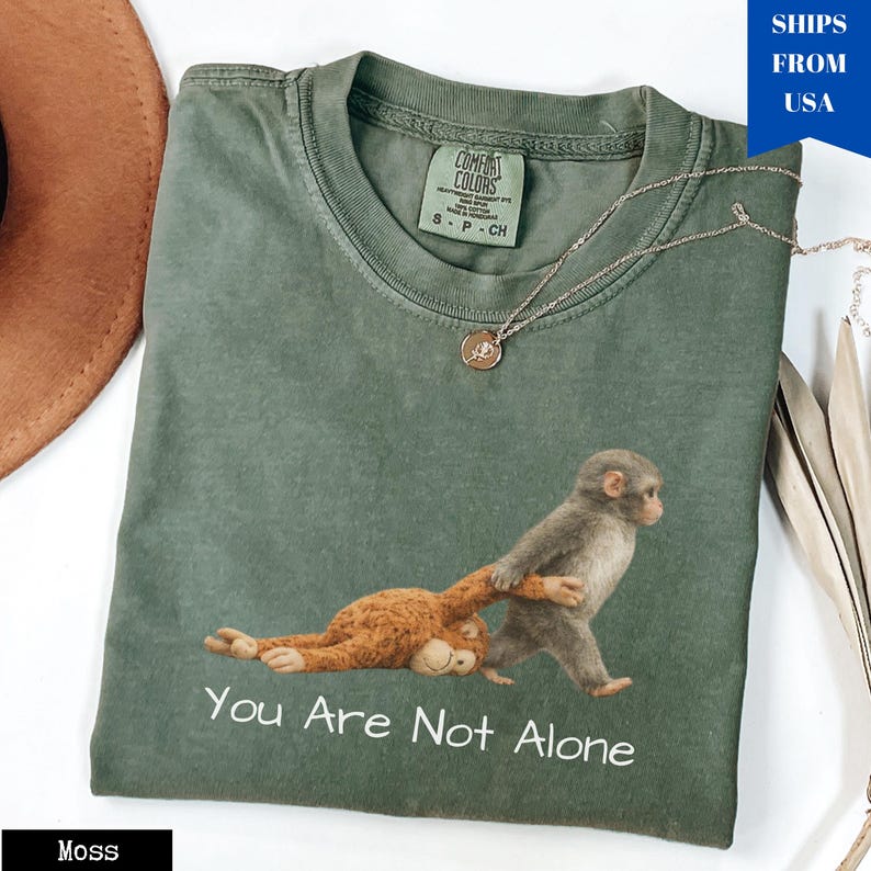 Puede incluir: Camiseta verde musgo con un gr&aacute;fico de monos y el texto "You Are Not Alone". Un collar plateado con un colgante est&aacute; sobre la camiseta. La camiseta tiene una etiqueta de Comfort Colors.