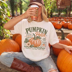 May include: An ivory short-sleeve t-shirt featuring a graphic print with the text PUMPKIN PATCH, FRESH FROM THE PATCH, FARM FRESH, LOCALLY GROWN, PUMPKINS, CORN MAZE, and APPLE CIDER, centred around an illustration of a large orange pumpkin.