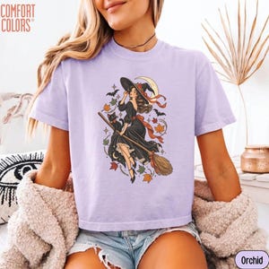 May include: A light purple short-sleeve t-shirt featuring a graphic print of a witch in a black dress riding a broomstick with a black cat, bats, and autumn leaves. The text COMFORT COLORS is printed in the top left corner of the shirt.