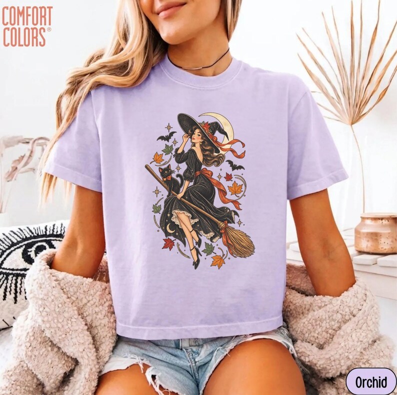 May include: A light purple short-sleeve t-shirt featuring a graphic print of a witch in a black dress riding a broomstick with a black cat, bats, and autumn leaves. The text COMFORT COLORS is printed in the top left corner of the shirt.