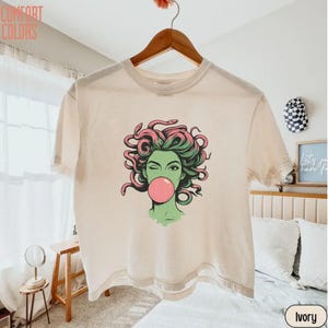 May include: Ivory t-shirt with a graphic of a woman with green skin, pink hair, and a bubblegum bubble. The design includes the text "Comfort Colors" at the top left. The shirt is hanging on a wooden hanger.