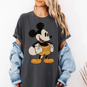 Könnte beinhalten: Dunkelgraues T-Shirt mit einem Vintage-Mickey-Mouse-Motiv. Die Cartoonfigur ist in Schwarz, Weiß und Gelb gehalten. Das T-Shirt hat kurze Ärmel und eine lockere Passform. Geeignet für Freizeitkleidung.