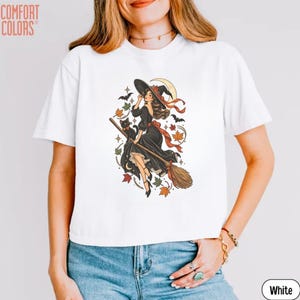 May include: A white t-shirt featuring a graphic print of a witch in a black dress riding a broomstick with a black cat, surrounded by autumn leaves, stars, a crescent moon, and bats. The text COMFORT COLORS is printed in the top left corner.
