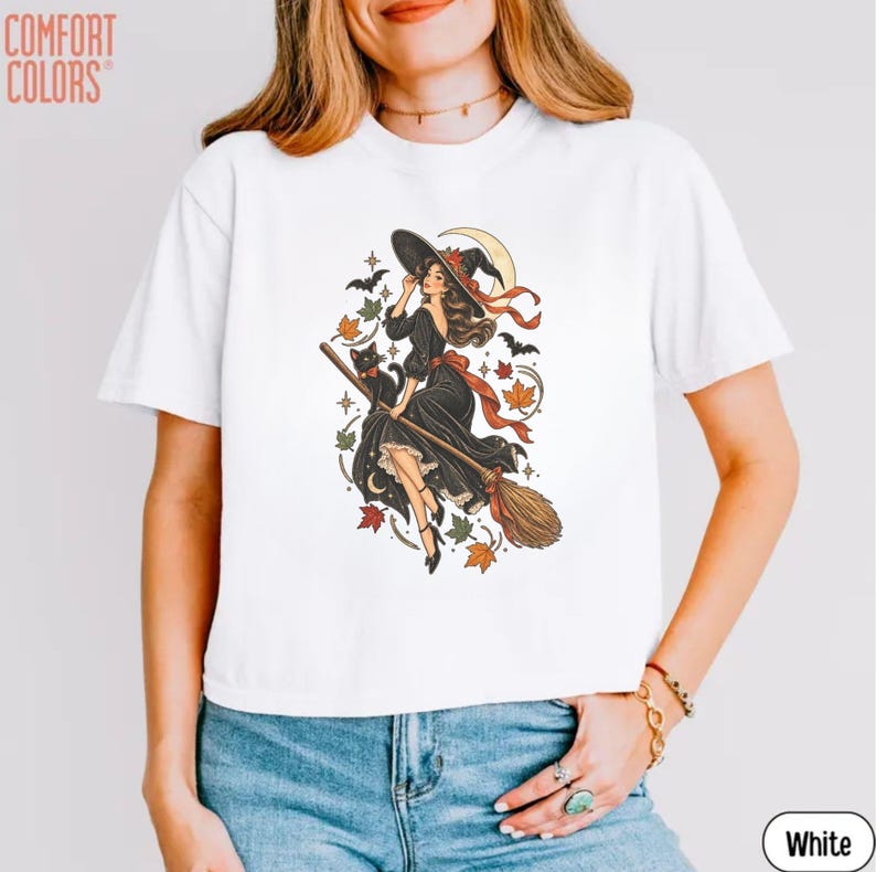 May include: A white t-shirt featuring a graphic print of a witch in a black dress riding a broomstick with a black cat, surrounded by autumn leaves, stars, a crescent moon, and bats. The text COMFORT COLORS is printed in the top left corner.
