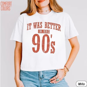 May include: A white short-sleeved t-shirt featuring the text IT WAS BETTER IN THE 90's in a distressed red font. The shirt has a crew neckline and is made of a solid white fabric. The Comfort Colors brand logo is visible in the top left corner.