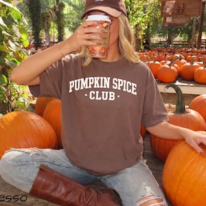 May include: A brown short-sleeved t-shirt with the white text PUMPKIN SPICE CLUB printed on the chest. The shirt is worn with light blue distressed denim jeans and brown boots, paired with a brown baseball cap and a clear plastic cup.
