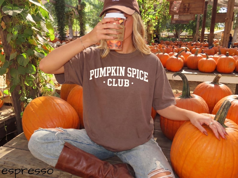 May include: A brown short-sleeved t-shirt with the white text PUMPKIN SPICE CLUB printed on the chest. The shirt is worn with light blue distressed denim jeans and brown boots, paired with a brown baseball cap and a clear plastic cup.
