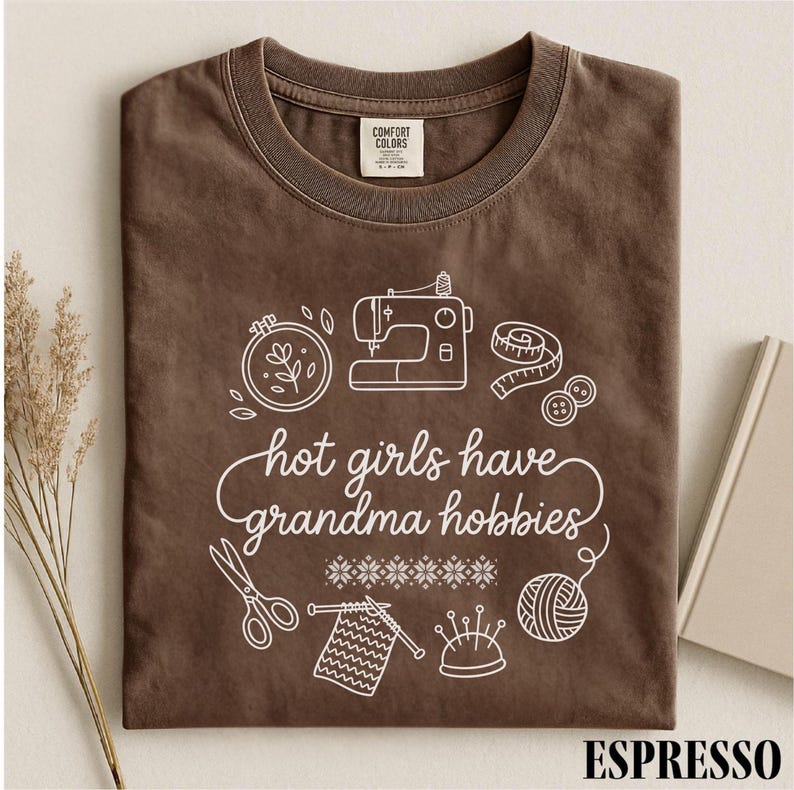 May include: Espresso-coloured t-shirt with a white graphic of sewing and knitting tools, and the phrase "hot girls have grandma hobbies." The shirt is folded and has a Comfort Colors label.
