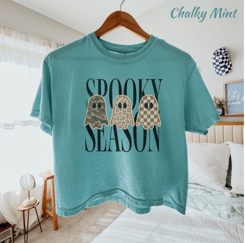 May include: A teal short-sleeve t-shirt featuring a graphic print of three ghosts with camouflage, leopard, and checkerboard patterns. The text "SPOOKY SEASON" is printed in black lettering behind the ghosts. The shirt is displayed on a wooden hanger.