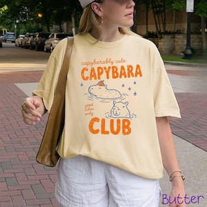 May include: A light yellow t-shirt featuring a graphic print with the text "capybarably cute CAPYBARA good vibes only CLUB". The design includes two illustrated capybaras in water, one with an orange fruit on its head, and blue stars.