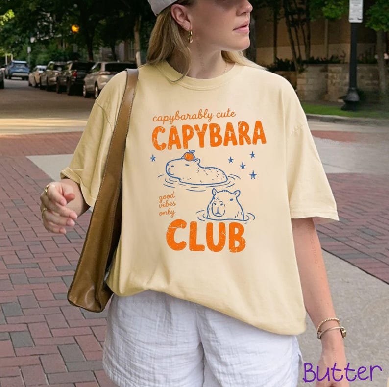 May include: A light yellow t-shirt featuring a graphic print with the text "capybarably cute CAPYBARA good vibes only CLUB". The design includes two illustrated capybaras in water, one with an orange fruit on its head, and blue stars.