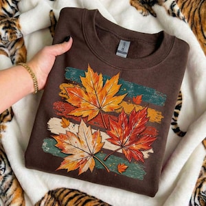 May include: A dark brown crewneck sweatshirt featuring a graphic print of three large maple leaves in shades of orange, yellow, and red. The leaves are layered over horizontal, distressed brushstroke stripes in teal, yellow, and cream colors.
