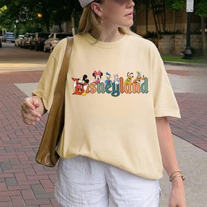 May include: A light yellow t-shirt featuring the word "Disneyland" in colorful, stylized lettering. The word is accompanied by cartoon character illustrations. The shirt has short sleeves and a relaxed fit.