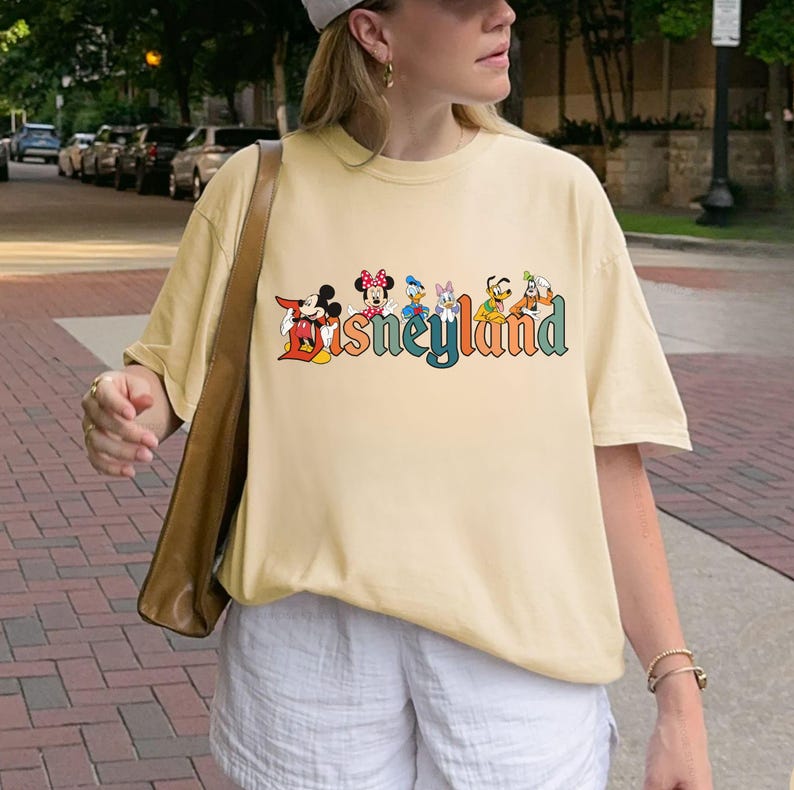 May include: A light yellow t-shirt featuring the word "Disneyland" in colorful, stylized lettering. The word is accompanied by cartoon character illustrations. The shirt has short sleeves and a relaxed fit.