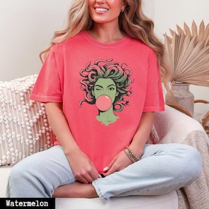 May include: A watermelon-coloured t-shirt with a graphic of Medusa winking and blowing a bubblegum bubble. The Medusa illustration has green hair and a green face. The word "Watermelon" is printed at the bottom of the image.